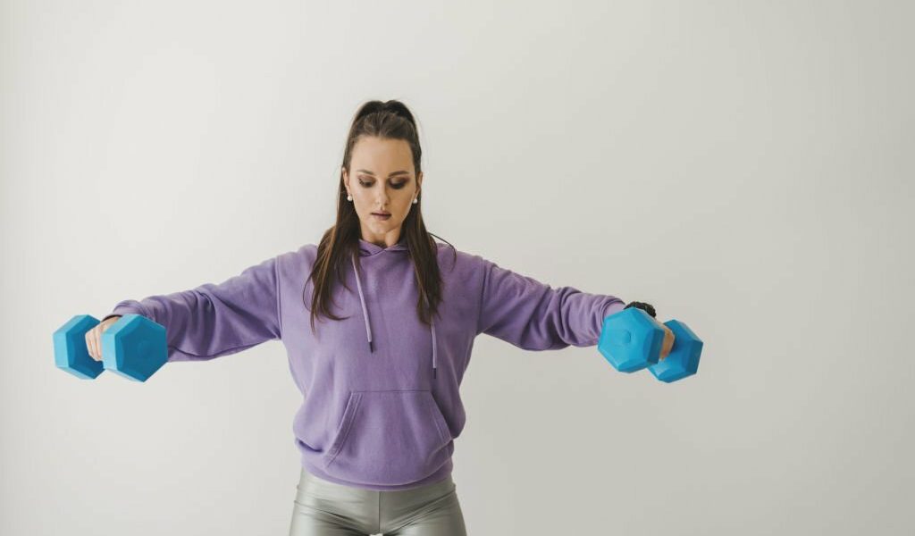 Young woman in purple hoodie and khaki shorts doing an indoor workout with dumbbells. Home based training with sport equipment