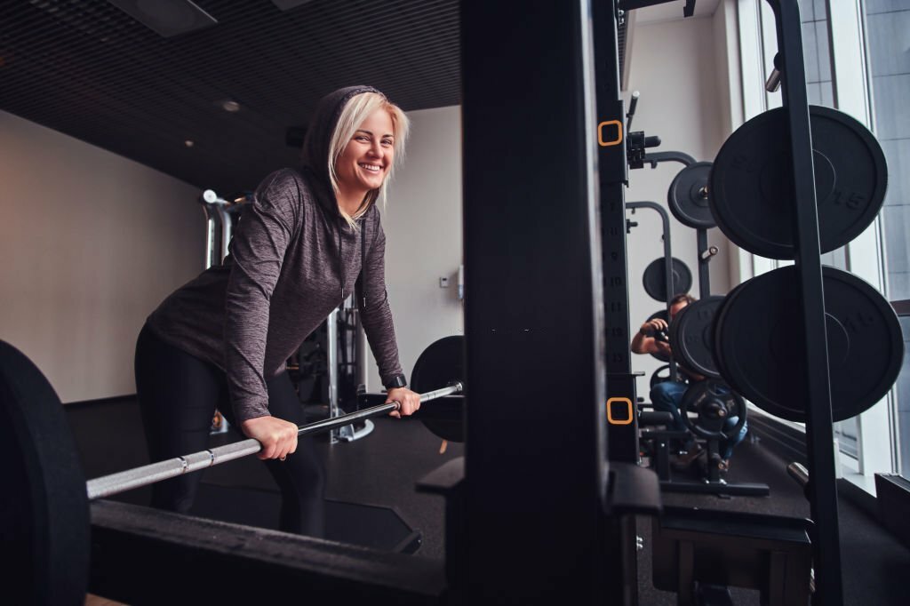 Beautiful sportive girl wearing hoodie leaning on a barbell next to a stand, smiling and looking at a camera