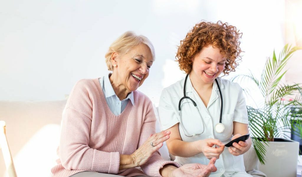 Happy senior woman having her blood sugar measured in a nursing home by her caregiver. Happy nurse measuring blood sugar of a senior woman in living room - diabetes